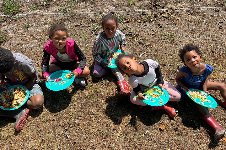 High-angle 45-degree shot of a diverse group of community students eating together at outdoor tables during an EQuivalence outreach day in Cape Town.