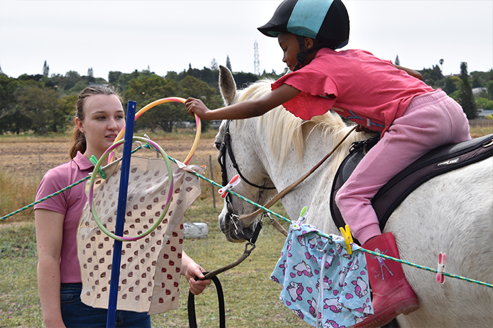 A student at EQuivalence in Durbanville practicing a coordination exercise by placing a large ring over a pole while mounted, assisted by a professional trainer.